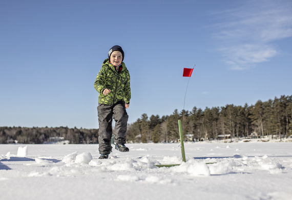 young boy in green snowsuit running to a red flag with blue skies and a snowy lake