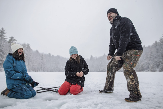 dad with two daughters laughing while sitting around an ice fishing trap on a lake on a snowy winter day