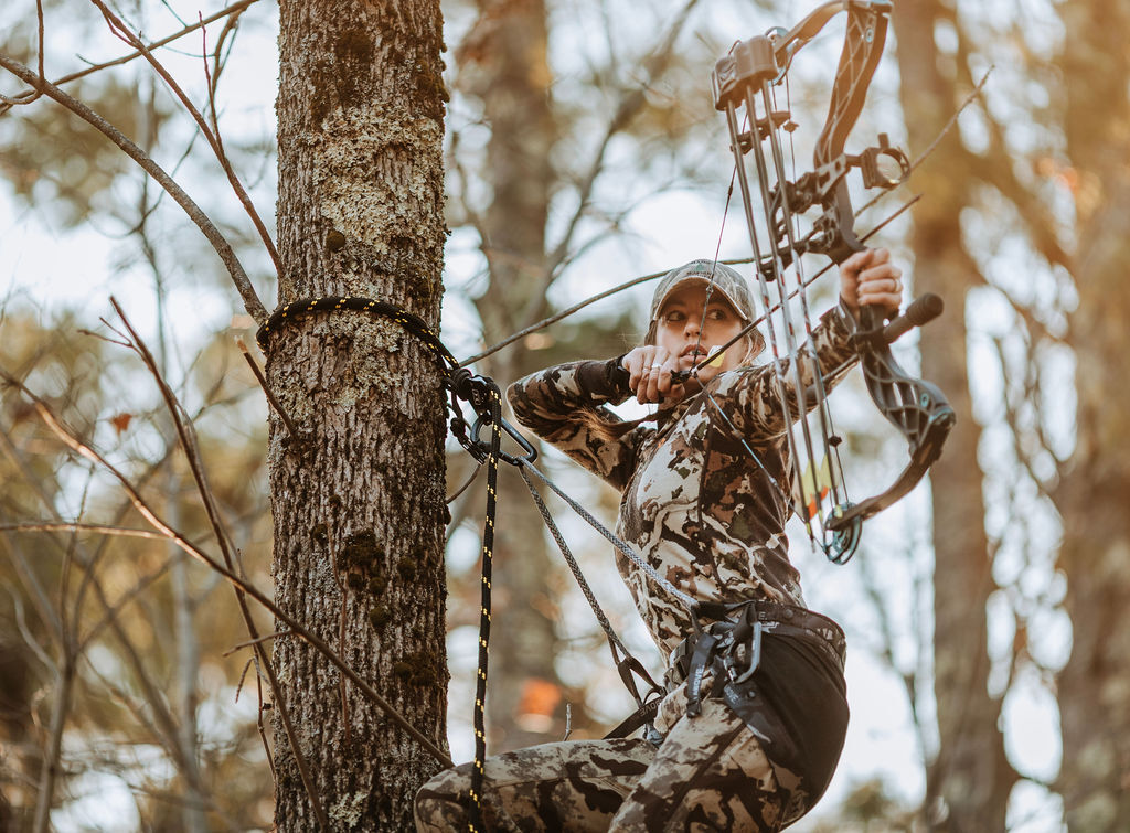 female archery hunting drawing back her bow wearing full camo in a saddle treestand