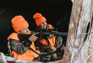 youth hunter aiming a firearm while in a blind with his mom wearing camo and blaze orange