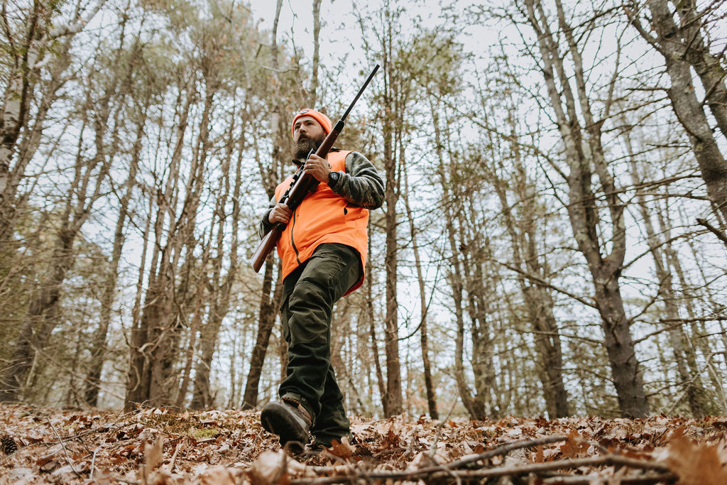 male hunter with large dark beard walking through woods with beard and camo with two articles of blaze orange