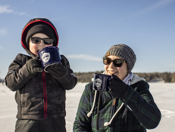 A kid sipping from a camp mug while ice fishing with his parent.