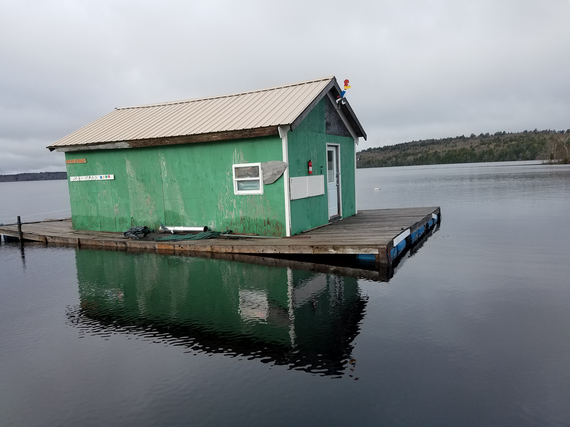 green stick built shack with roof and a door and one window on a floating dock in middle of a lake