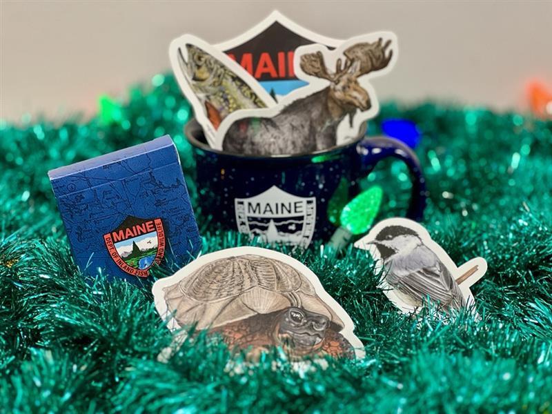 deck of cards, a MDIFW blue mug with white logo, and sticker decals of a moose wood turtle trout and MDIFW logo on a decorative holiday background