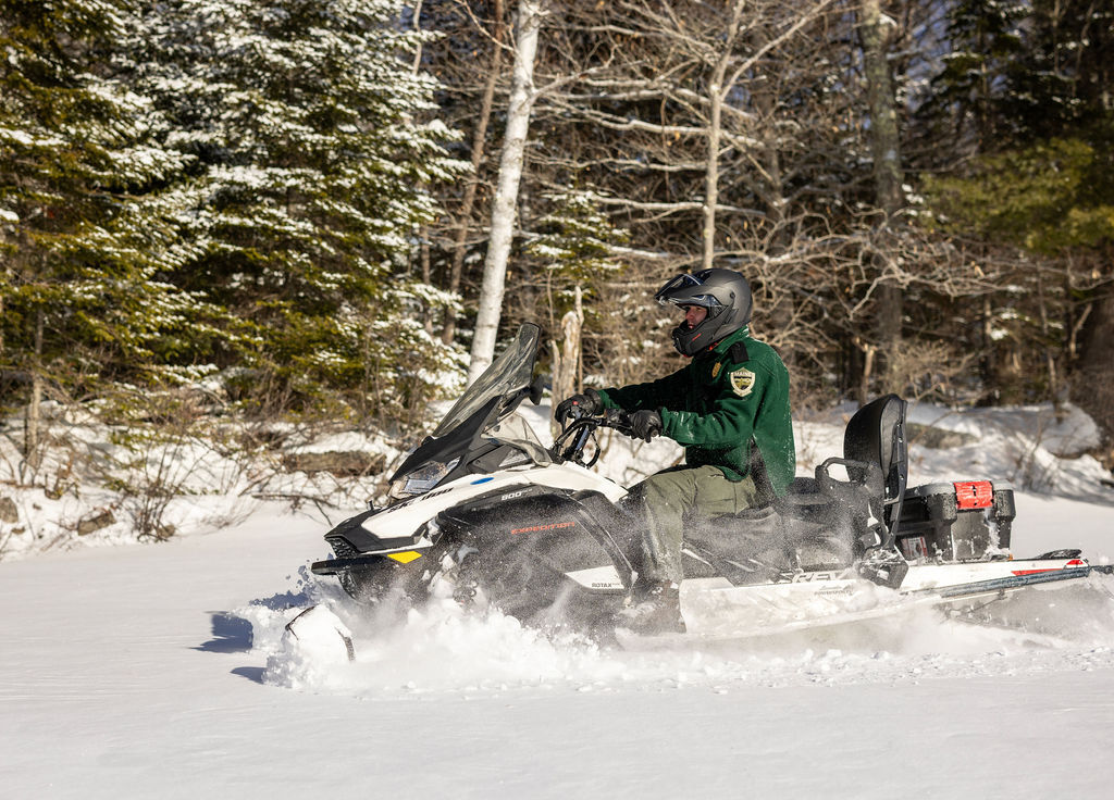 Game warden wearing green jacket with Warden Service patch on sleeve driving a new snowmobile through fresh snow on a lake