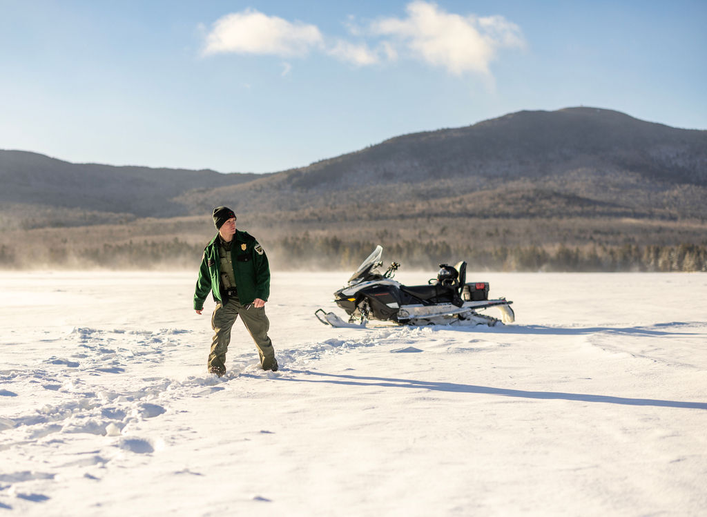 warden wearing green uniform walking from snowmobile to talk with an angler with mountain range backdrop and sunny skies