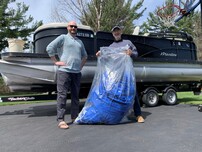 Two men stand in front of a boat with a bag of shrink wrap, ready to be recycled.