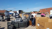A picture of piles of televisions and other electronic equipment in a large parking lot behind a brick building.
