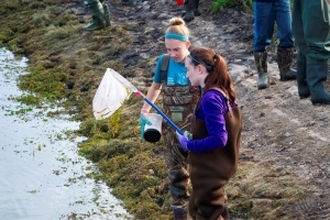 Two female students with nets stand by shore of pond