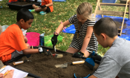Three elementary school children use garden tools and brushes in a container of dirt.