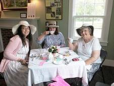 Three ladies in dresses and hats sit at a table set with a formal tea service.