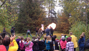 A group of schoolchildren watch three men dressed in Civil War uniforms fire a large cannon. 