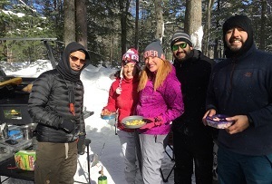 Campers dressed in winter gear, holding breakfast plates, in a snowy forest