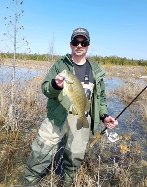 A man dressed in fishing gear, sunglasses and baseball cap, standing in water holding a fishing pole and a caught fish