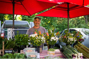 A farmer smiles at the camera from behind a table filled with flowers and vegetables.