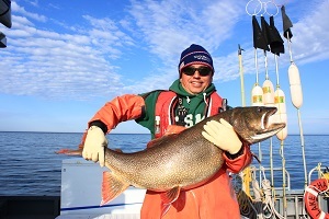 DNR fisheries biologist in a hat and sunglasses holds a redfin lake trout while standing on a boat deck, blue sky in background