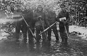 A group of seven men from CCC Camp Harrison, Company 1613 in Clare County, stand ankle deep in a river while building a bridge. 
