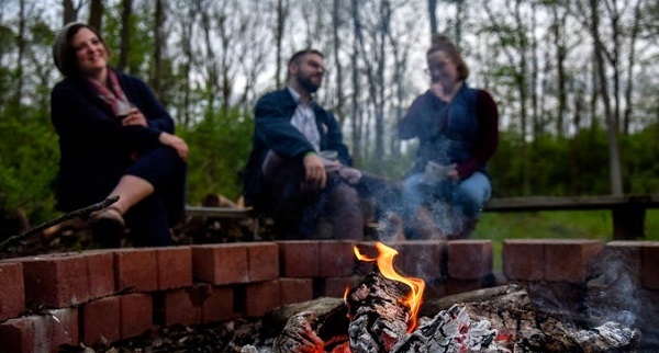 Two women and a man sitting on benches, campfire in the foreground, dusky forest in the background