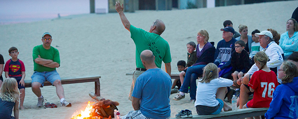 park staffer points to sky as visitors seated around campfire listen