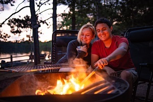 Woman and man sitting together around a campfire, woods in the background