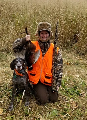 A smiling young hunter, dressed in camouflage and hunter orange, holding a pheasant by the feet, black hound at his side