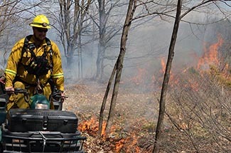 A Michigan DNR firefighter rides an off-road vehicle past some burning bushes.