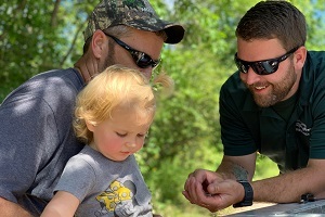 Two men, both wearing sunglasses and one holding a little girl, look at a juvenile lake sturgeon in the other man's hands