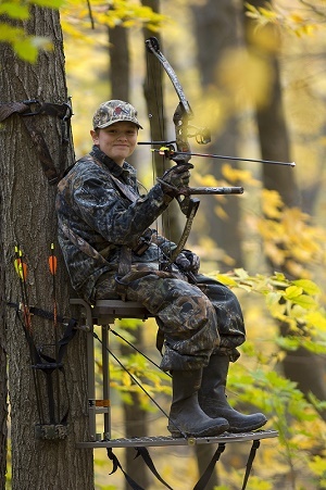 A young man wearing camo and using a full-body harness, bow hunting from a tree stand