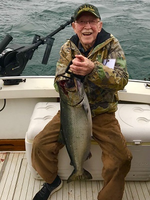 A smiling, older man dressed in camo, wearing a Navy hat, holding a chinook salmon while sitting on a cooler on a boat on Lake Michigan