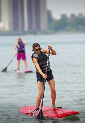 Two women stand-up paddle boarding on the Detroit River, with downtown office buildings in background