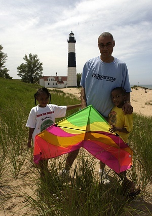 An African American man and two small children holding a multicolored kite on the beach in front of Big Sable Point Lighthouse