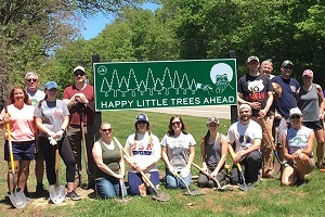 Smiling volunteers holding shovels and surrounding a Happy Little Trees Ahead sign, bearing the likeness of Bob Ross