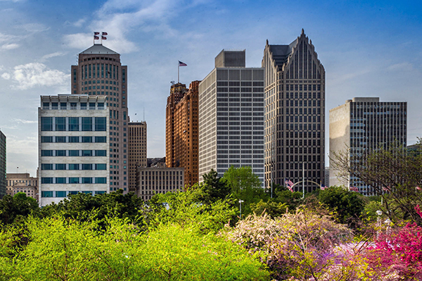 The skyline of Detroit is shown with green and flowering trees in the foreground.