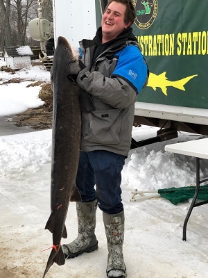 Smiling man wearing winter gear holds up a large sturgeon; standing in front of DNR registration station on Black Lake