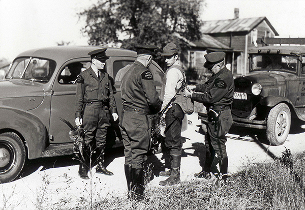 A historic photo shows conservation officers with a hunter stopped along a roadside, checking his license and bag.