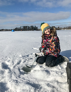 A young girl is shown sitting on the ice with a scoop she uses to clear ice from her fishing hole.
