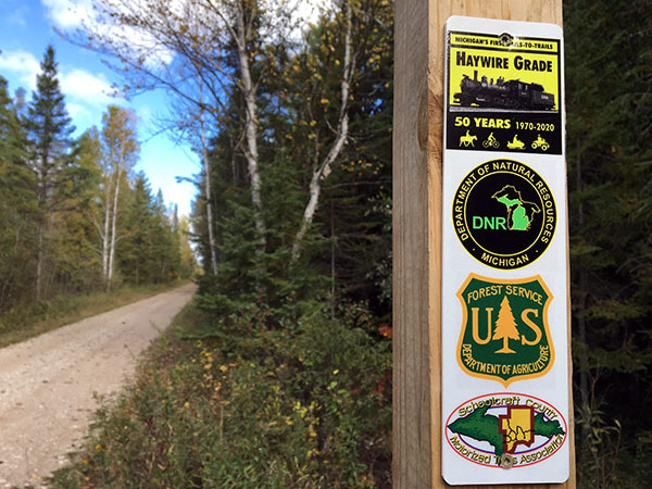 A trail marker along the Haywire Grade shows logos for the 50th anniversary celebration and partner organizations.