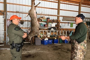 A DNR conservation officer talks with a male hunter, in a pole barn, with a harvested deer hanging in the background