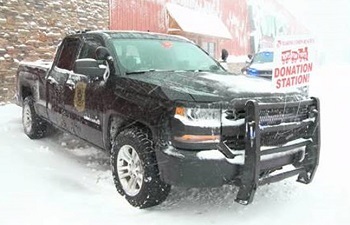 front view of a parked, black DNR conservation officer patrol truck, with a nearby holiday donations sign, while snow is falling