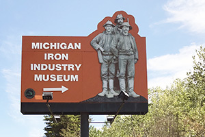 A billboard features a bright orange background, a black and white photo of miners, and the words "Michigan Iron Industry Museum."