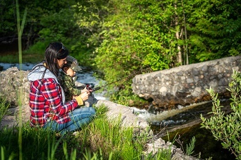 a woman with long dark hair, wearing a red plaid jacket, helps a young boy wearing a baseball cap hold a fishing pole out over a grassy, rocky ridge