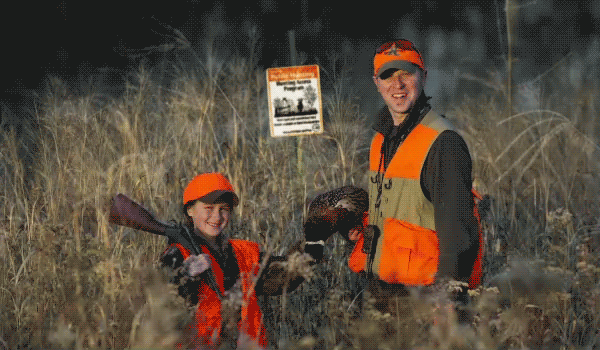A father and his daughter wearing hunter orange, holding harvested pheasants, stand in a grassy field in front of a Hunter Access Program sign.