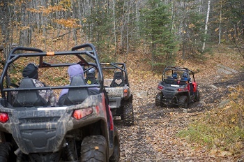 three black and camo all-terrain vehicles driving away single file, down a muddy leaf-covered forest road