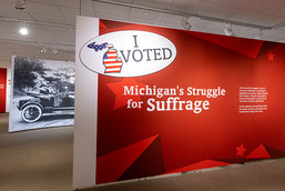 A wide angle view of the entrance to a gallery includes a red wall with the text "I Voted" and a large image of a historic car.