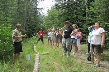 A man in DNR short and shorts talks to a group of male and female conference attendees in the woods