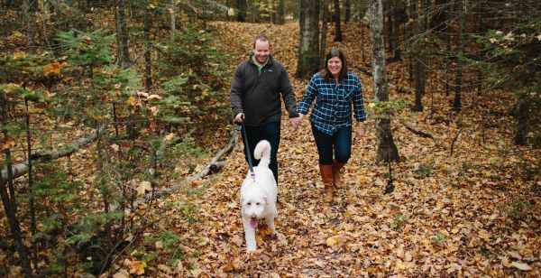 A couple wearing sweaters walks a fluffy white dog on a trail covered in autumn leaves 