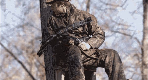 A man wearing camouflage clothing sits in a tree stand holding a crossbow looking over his shoulder while the camera zooms out.