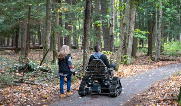 A woman and a man using a track chair enjoy a paved path in autumn