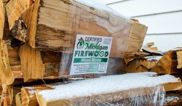 A bundle of firewood with a "certified Michigan firewood" tag. The bundle is outdoors, and a dusting of snow is on the plastic wrapper.