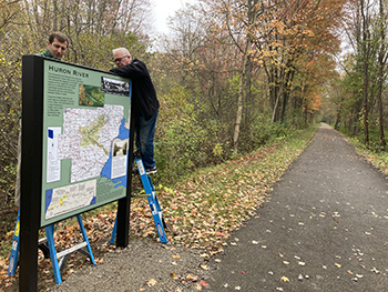 Two men on ladders use tools to fasten a 3' x 4' sign into a metal frame along a trail. 